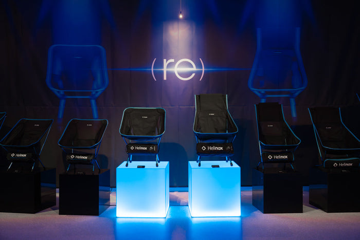 Lineup of black Helinox camp chairs with blue trim displayed on pedestals in front of a (re) backdrop
