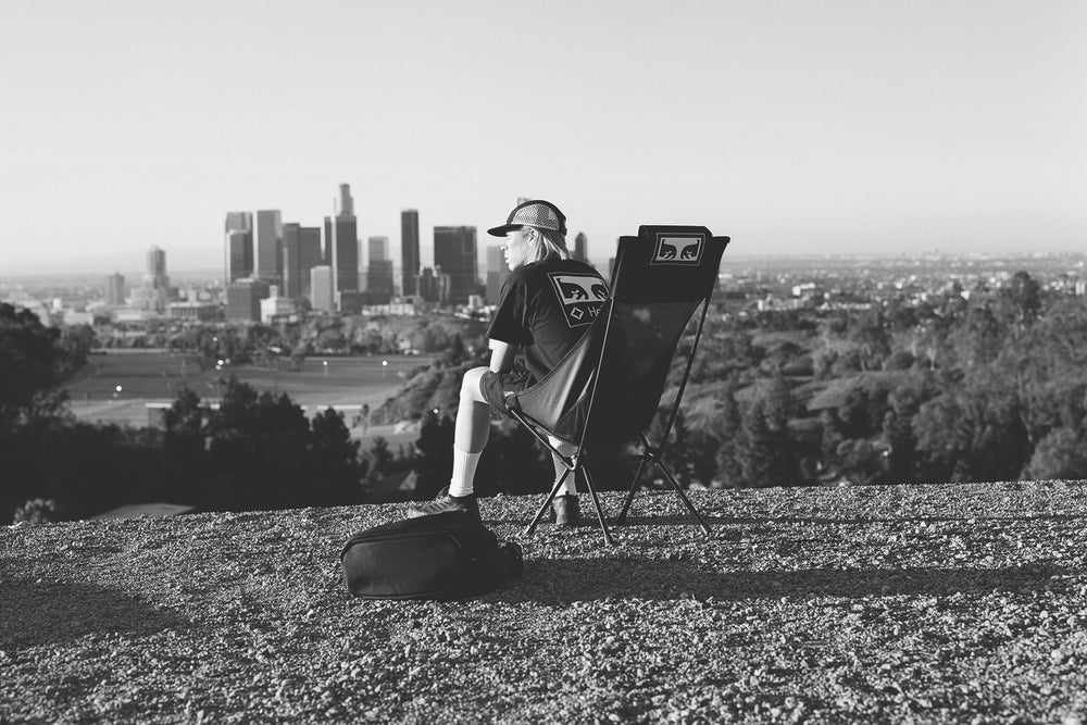 A man sitting in a Helinox x Obey high-back camp chair overlooking the Los Angeles city skyline
